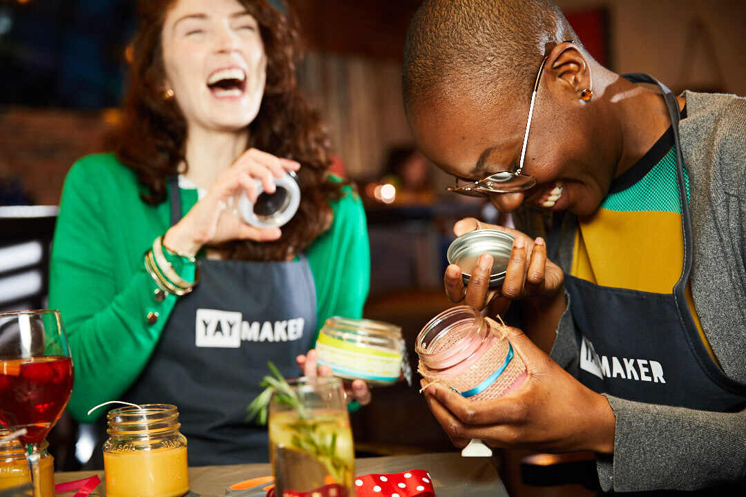 Two women laughing in Yaymaker aprons holding hand crafted candles